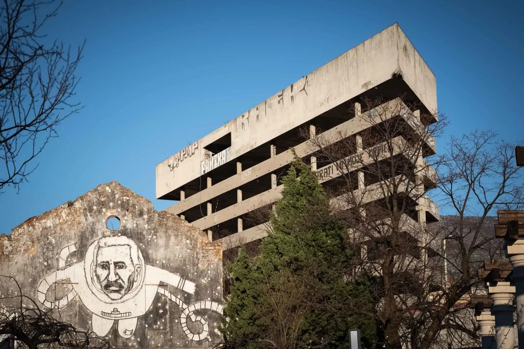 Zrujnowany betonowy budynek z graffiti i zniszczona ściana z dużym muralem przedstawiającym mężczyznę w skafandrze, Mostar.