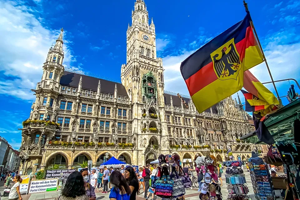 The ornate facade and clock tower of Munich's New Town Hall under a blue sky, with a German flag and market stalls.