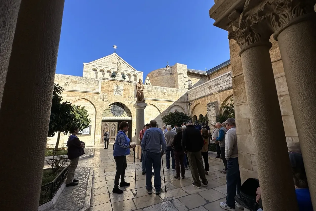 Turistas en un patio de piedra, frente a la fachada de una iglesia histórica con arcos, una estatua y una cruz, bajo cielo...