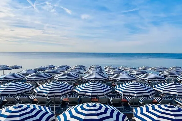Rzędy niebiesko-białych parasoli plażowych i leżaków na kamienistej plaży, z morzem i błękitnym niebem w tle.