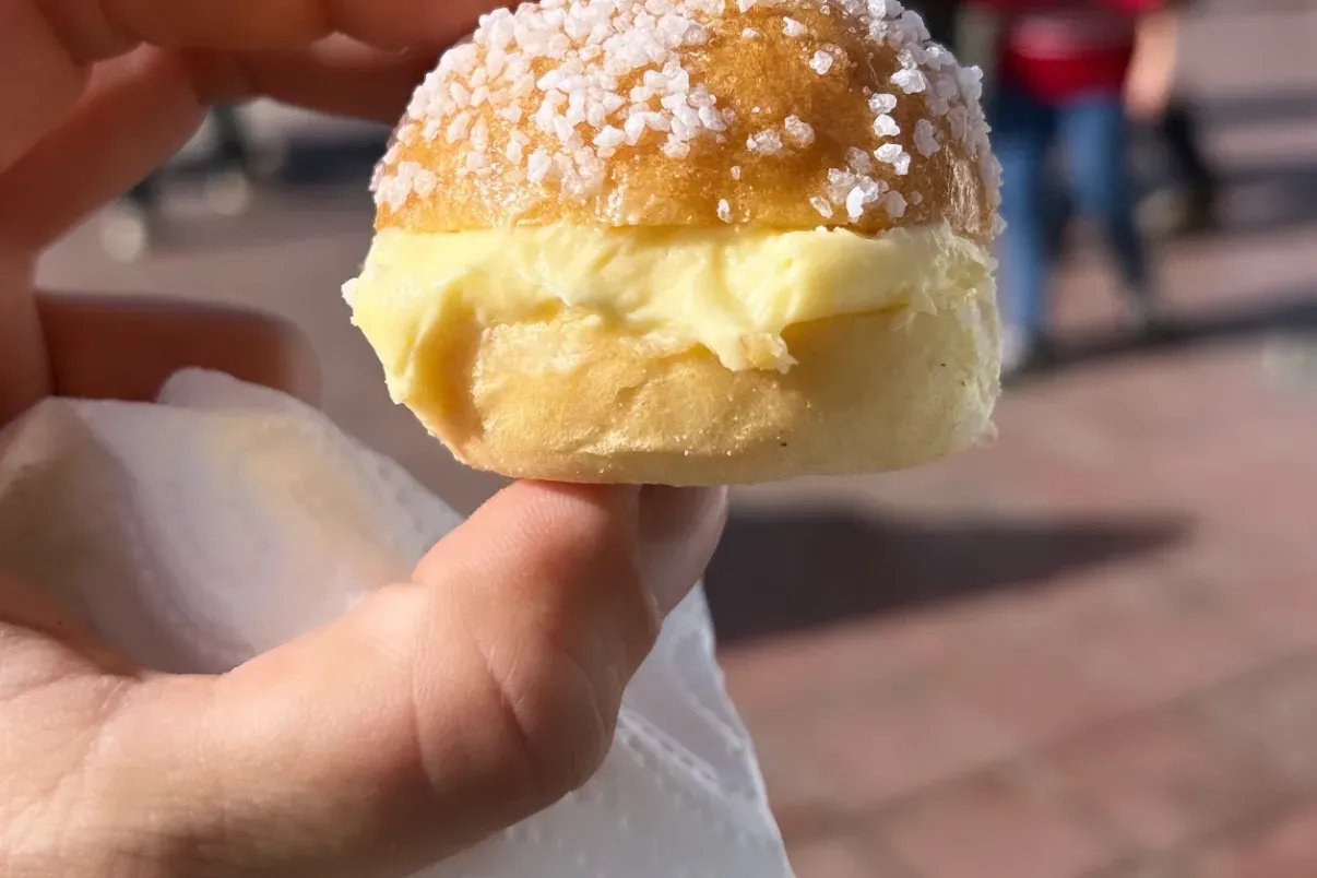 Hand hält ein kleines, briocheartiges Gebäck mit Cremefüllung und grobem Zucker. Im Hintergrund sind Menschen und Pflaster...