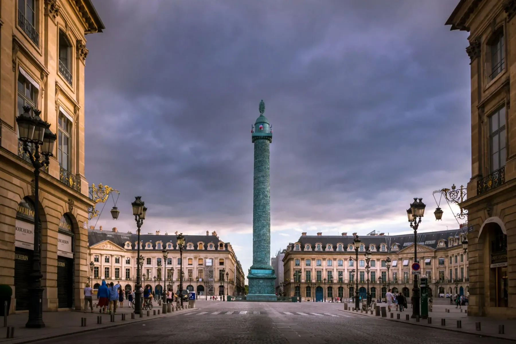 Die Vendôme-Säule auf dem Place Vendôme in Paris, umrahmt von eleganten Gebäuden unter bewölktem Himmel. Passanten auf Kop...