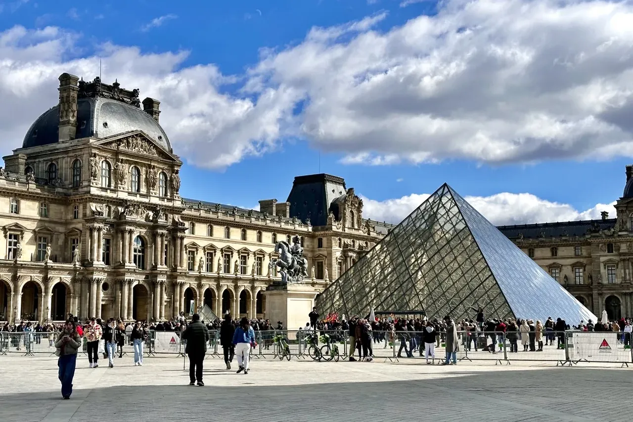 Blick auf das Louvre-Museum mit klassischer Architektur und Glaspyramide, unter teils bewölktem blauem Himmel. Personen im...