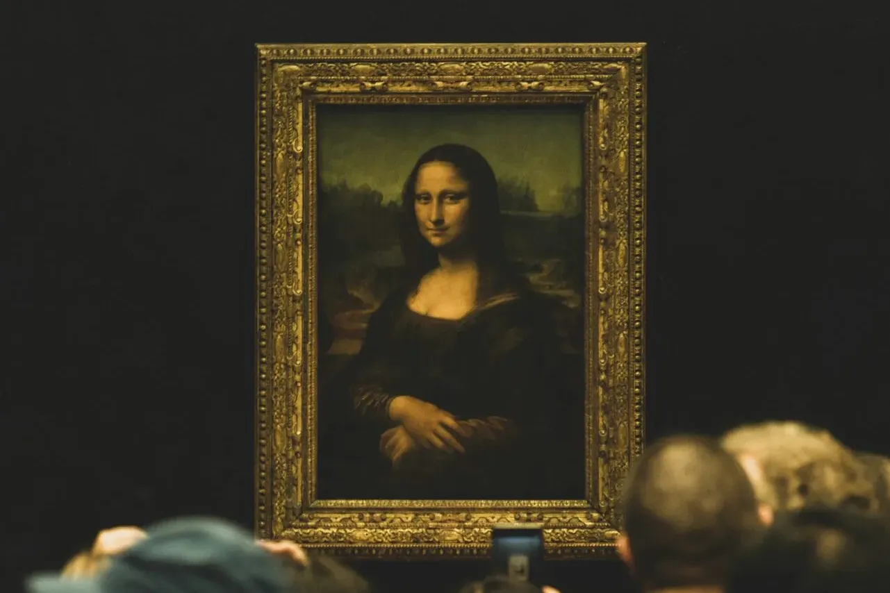 Das Gemälde der Mona Lisa in einem verzierten Rahmen, an einer dunklen Wand, mit unscharfen Besuchern im Vordergrund.