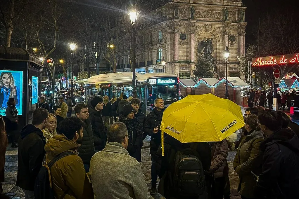 Walkative-Reisegruppe nachts, versammelt am beleuchteten Molière-Brunnen in Paris, mit Weihnachtsmarktständen.
