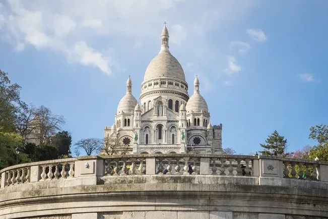 Die weiße Basilika Sacré-Cœur in Paris mit Kuppeln, von unterhalb einer geschwungenen Steinbalustrade unter blauem Himmel ...