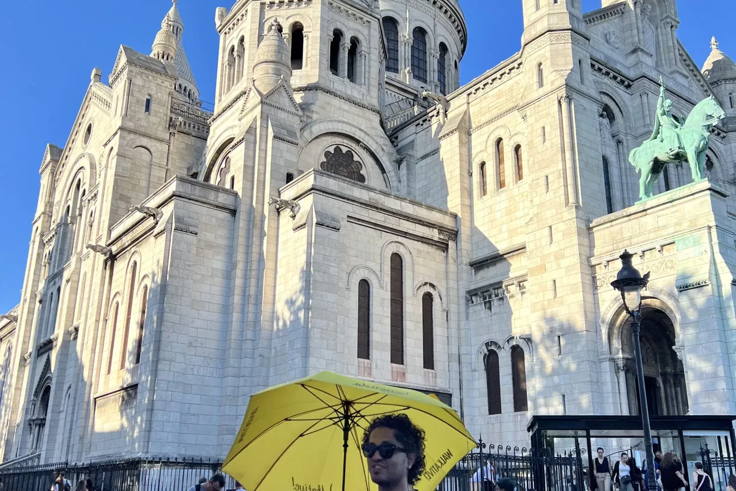 Weiße Kuppeln und Fassade der Sacré-Cœur-Basilika unter blauem Himmel. Eine Person mit gelbem Regenschirm auf Kopfsteinpfl...