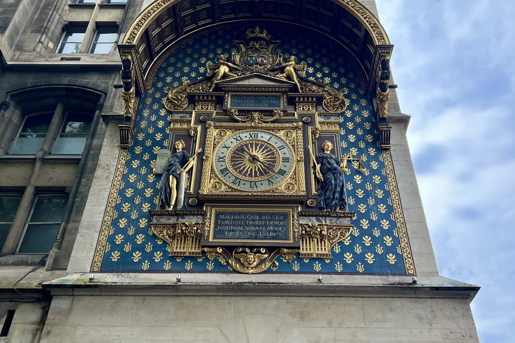 Verzierter goldener Uhrenschild auf historischem Steinturm mit blau-goldenem Fleur-de-lis-Muster, flankiert von zwei Statu...