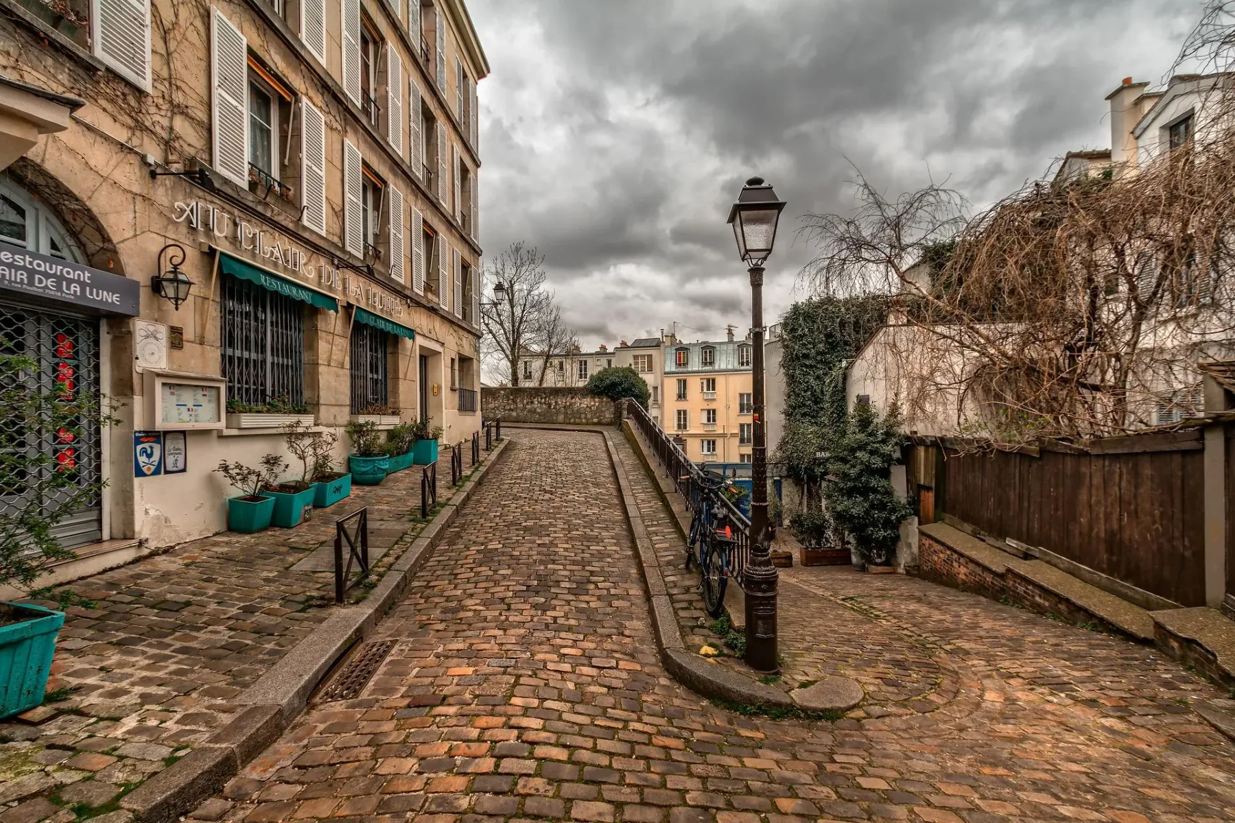 Gewundene Kopfsteinpflasterstraße in Paris, mit einem Steinhaus und Restaurant links, und einer Straßenlaterne rechts.