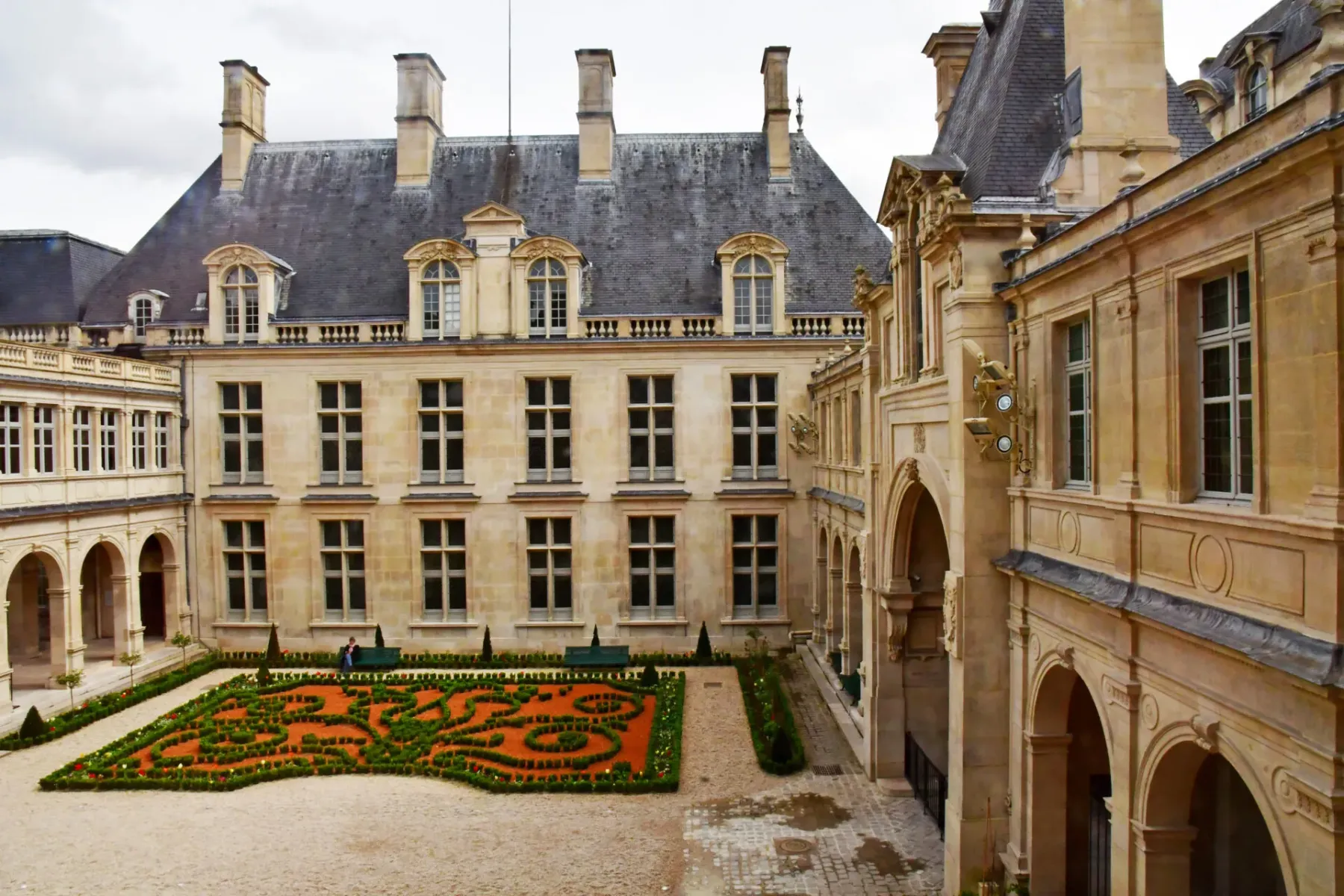 Ein großer Pariser Innenhof mit einem kunstvoll geformten Ziergarten, umgeben von hellen Steingebäuden mit Gaubenfenstern.