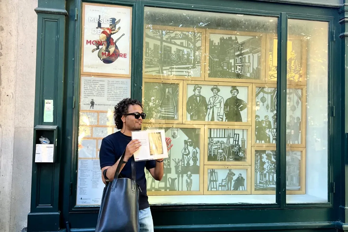 Mann mit Sonnenbrille und Buch vor Le Bateau-Lavoir, mit historischen Montmartre-Illustrationen in den Fenstern.