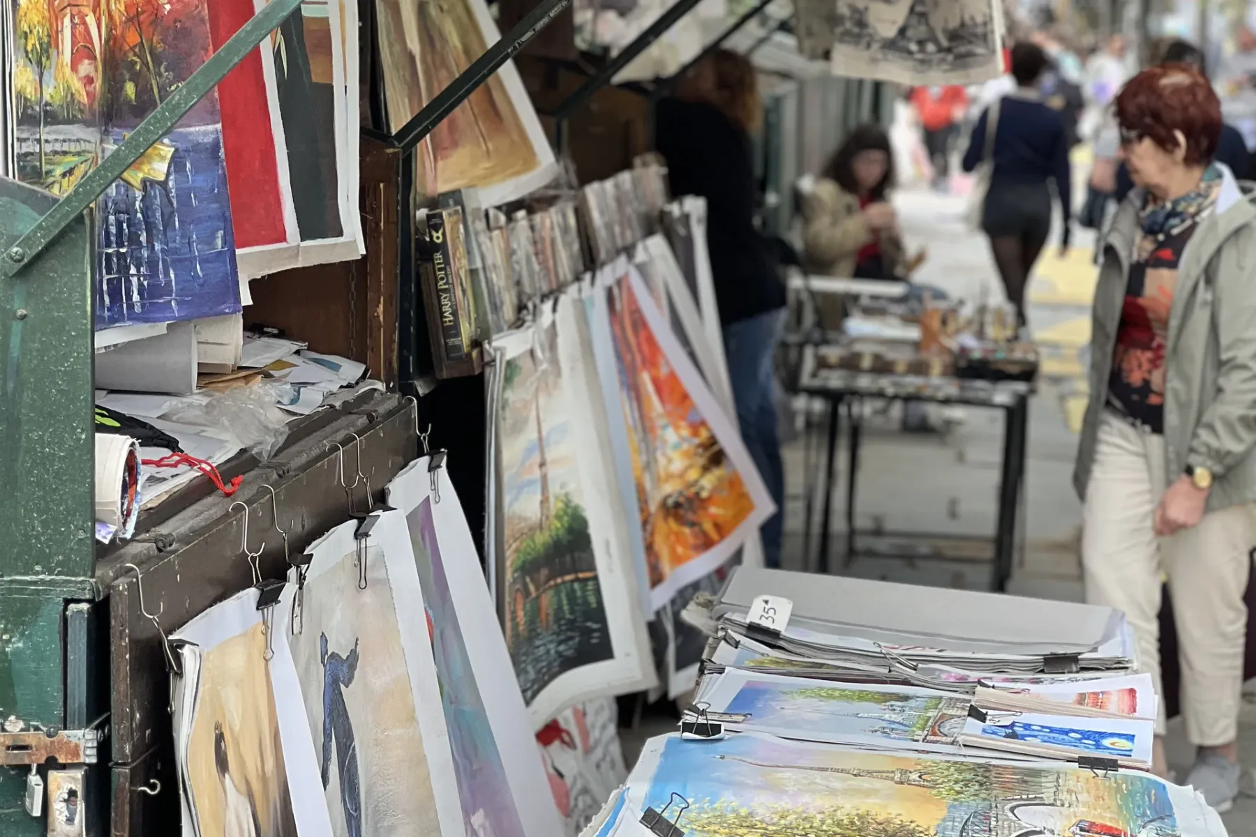 Street stall in Paris displaying various paintings and prints, including Eiffel Tower scenes and abstract art, with people...