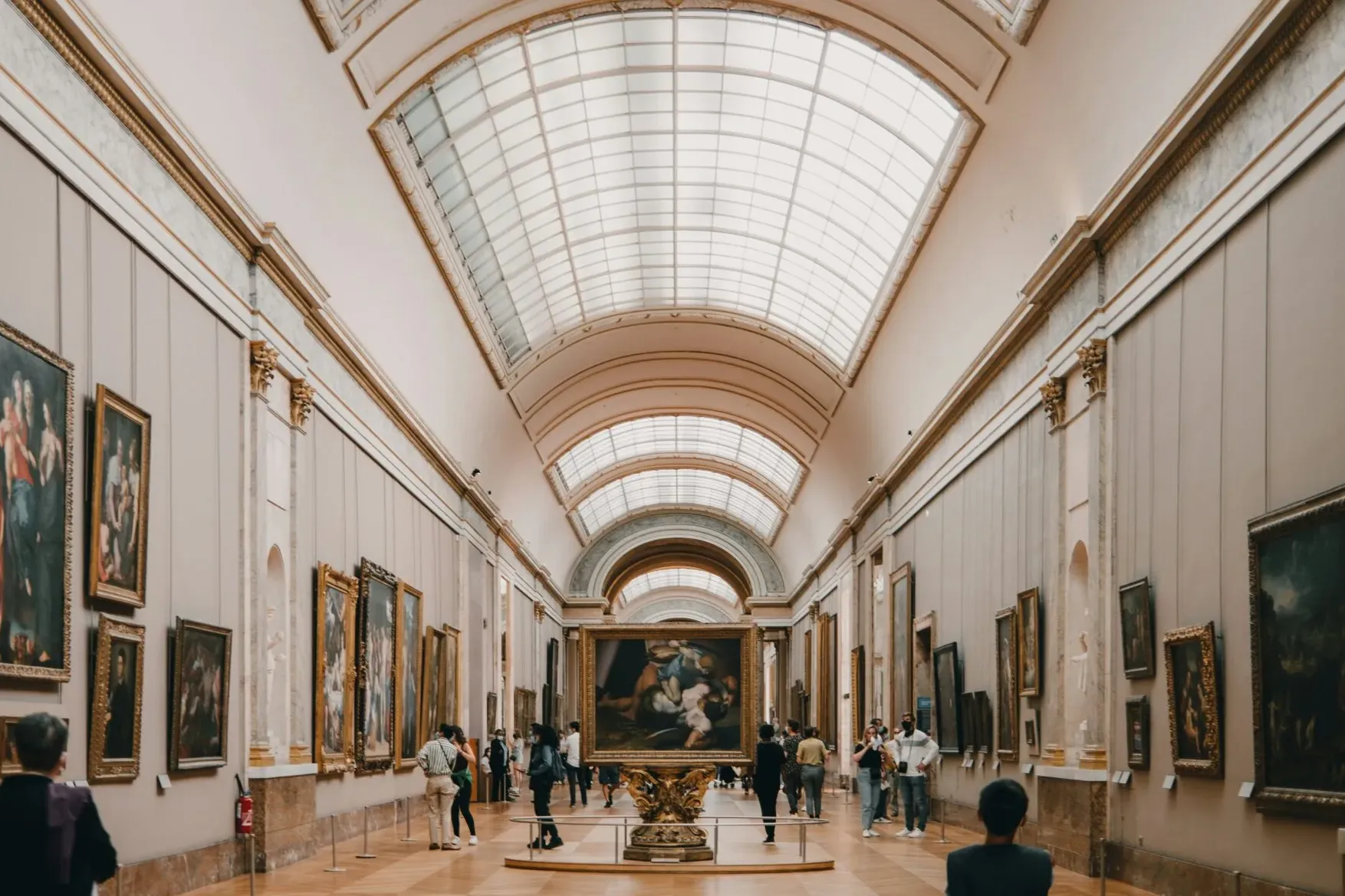 Große Kunstgalerie in Paris mit zahlreichen Gemälden an den Wänden und einer gewölbten Glasdecke. Besucher betrachten die ...