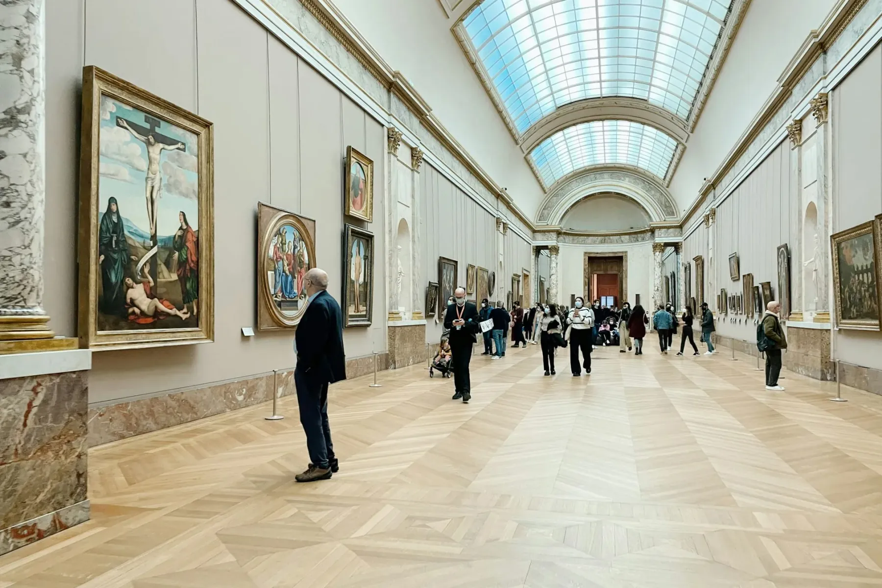 Lange Museumsgalerie mit Holzparkettboden, Gemälden an hellen Wänden und mehreren Besuchern. Ein großes Oberlicht erhellt ...