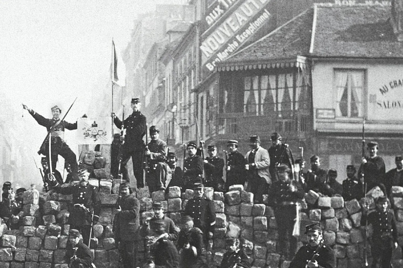 Schwarz-Weiß-Foto von Männern in Militäruniformen auf einer Kopfsteinpflaster-Barrikade in einer Pariser Straße mit Gebäuden.