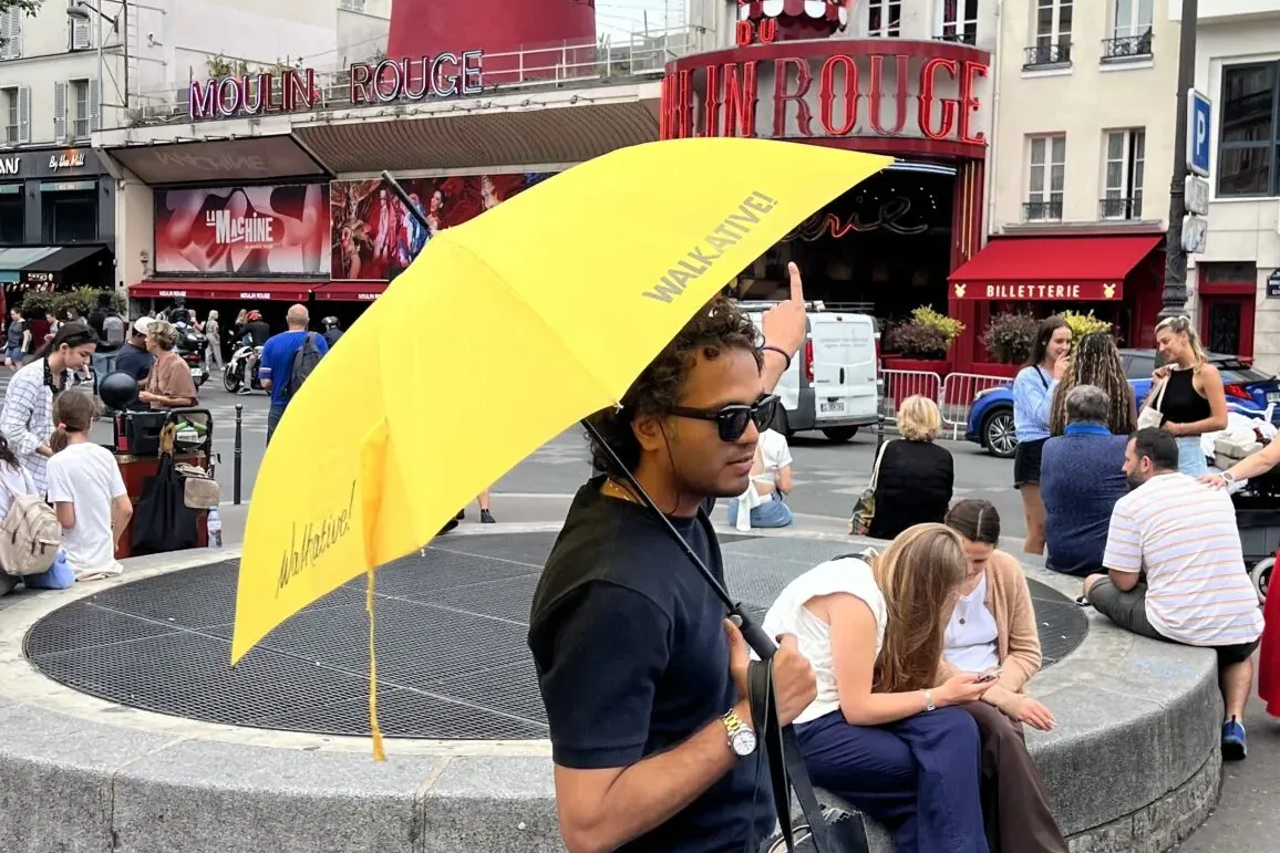 Ein Mann mit Sonnenbrille und gelbem Walkative-Regenschirm, der nach oben zeigt, mit dem roten Moulin Rouge Gebäude in Par...