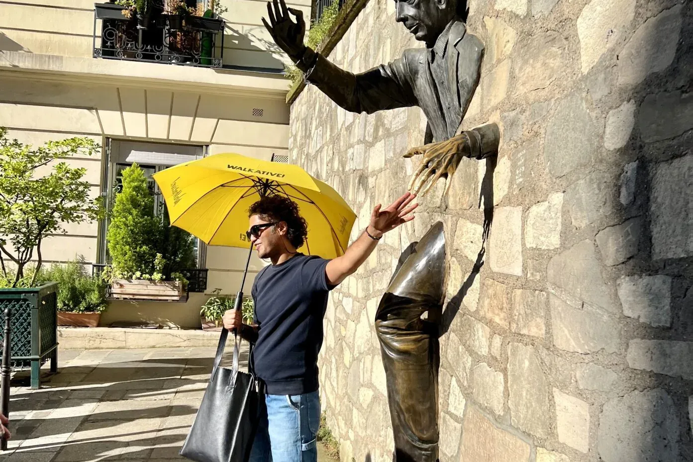Mann mit gelbem Walkative-Regenschirm berührt eine Bronzeskulptur eines Mannes, der aus einer Steinmauer tritt.