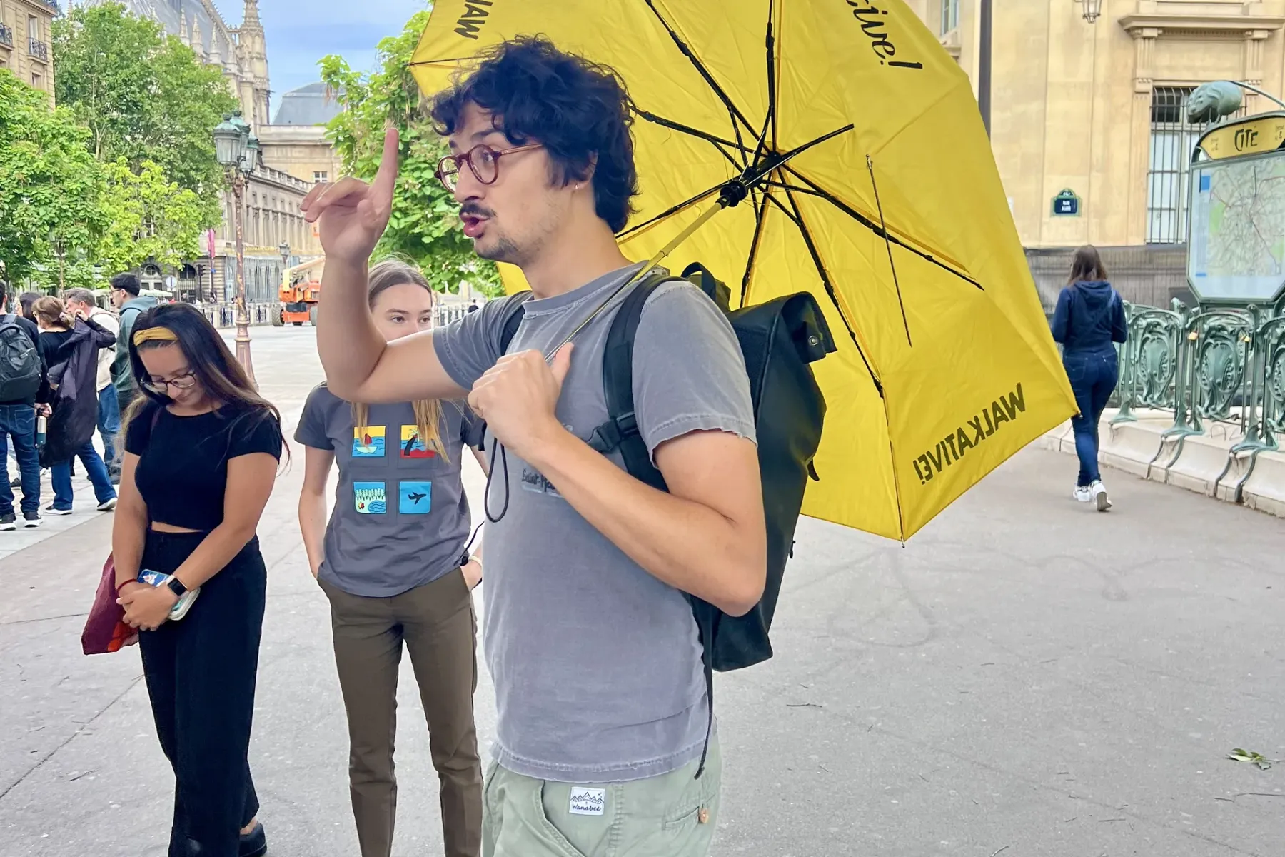 Male guide with yellow Walkative umbrella talking to a group on a Paris street, Sainte-Chapelle spire in background.