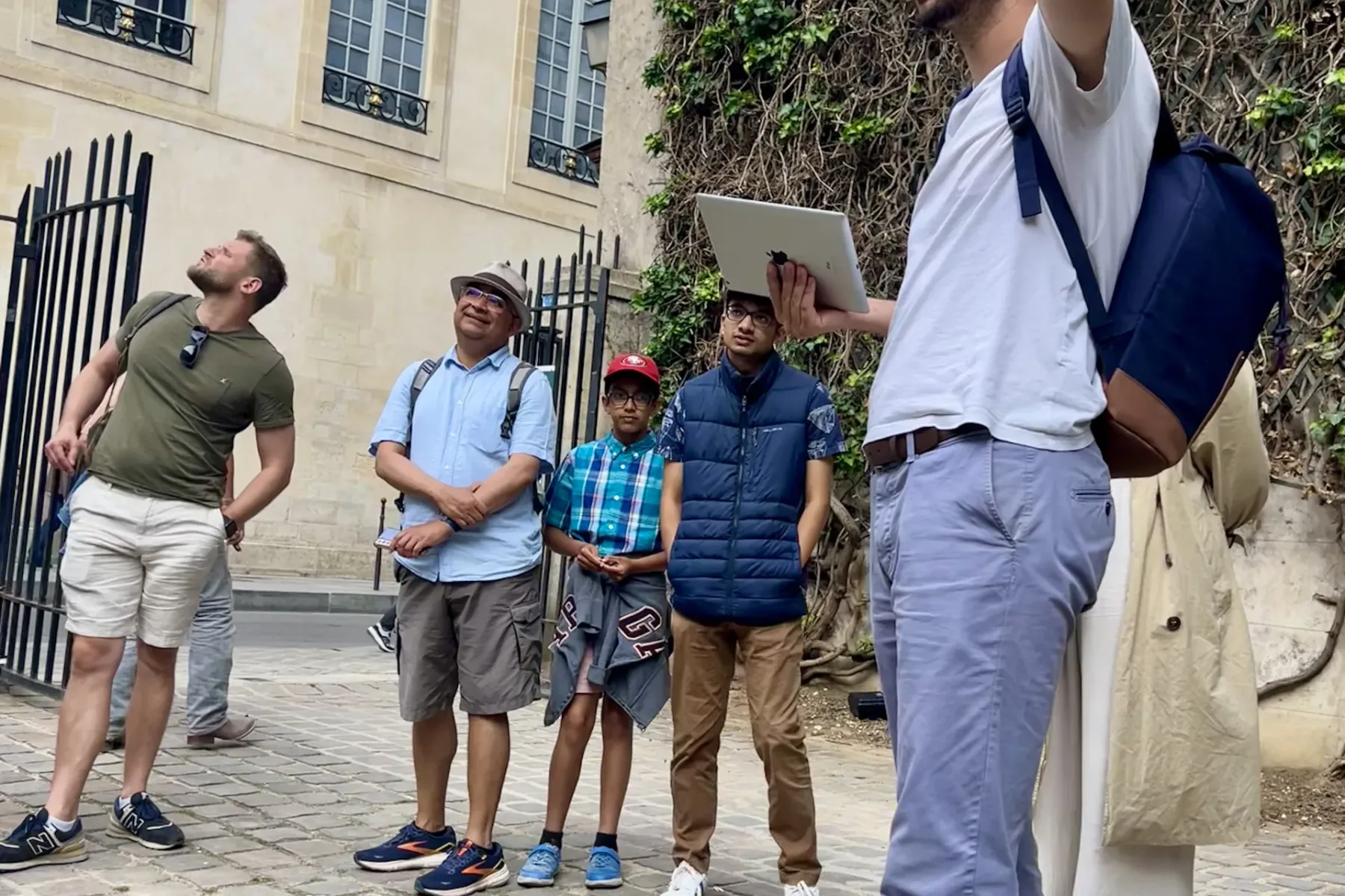 Reiseführer mit Tablet gestikuliert vor einem Steingebäude mit Efeu zu einer Gruppe von fünf Touristen auf Kopfsteinpflaster.