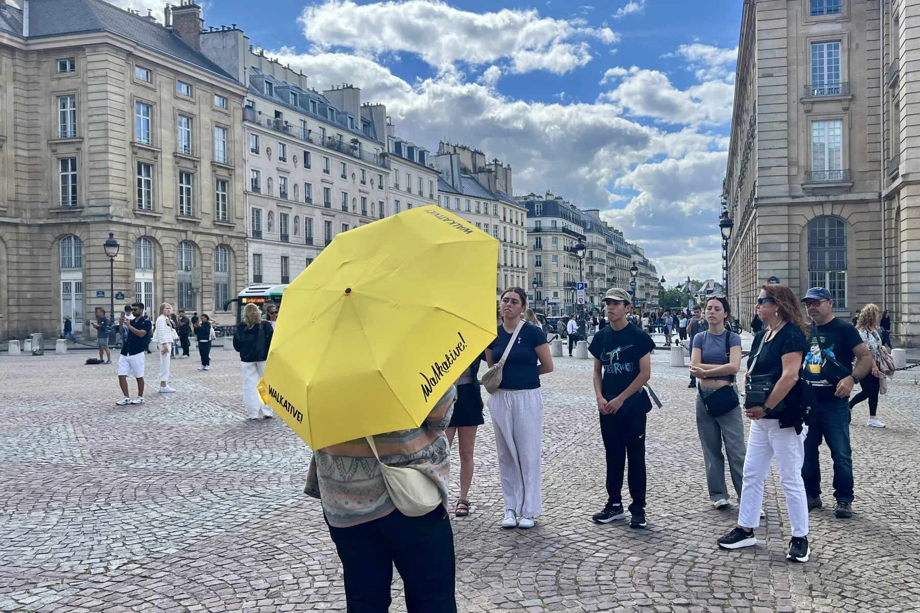 Rückenansicht einer Person mit gelbem Walkative-Regenschirm auf einem Kopfsteinpflasterplatz in Paris. Im Hintergrund Pari...