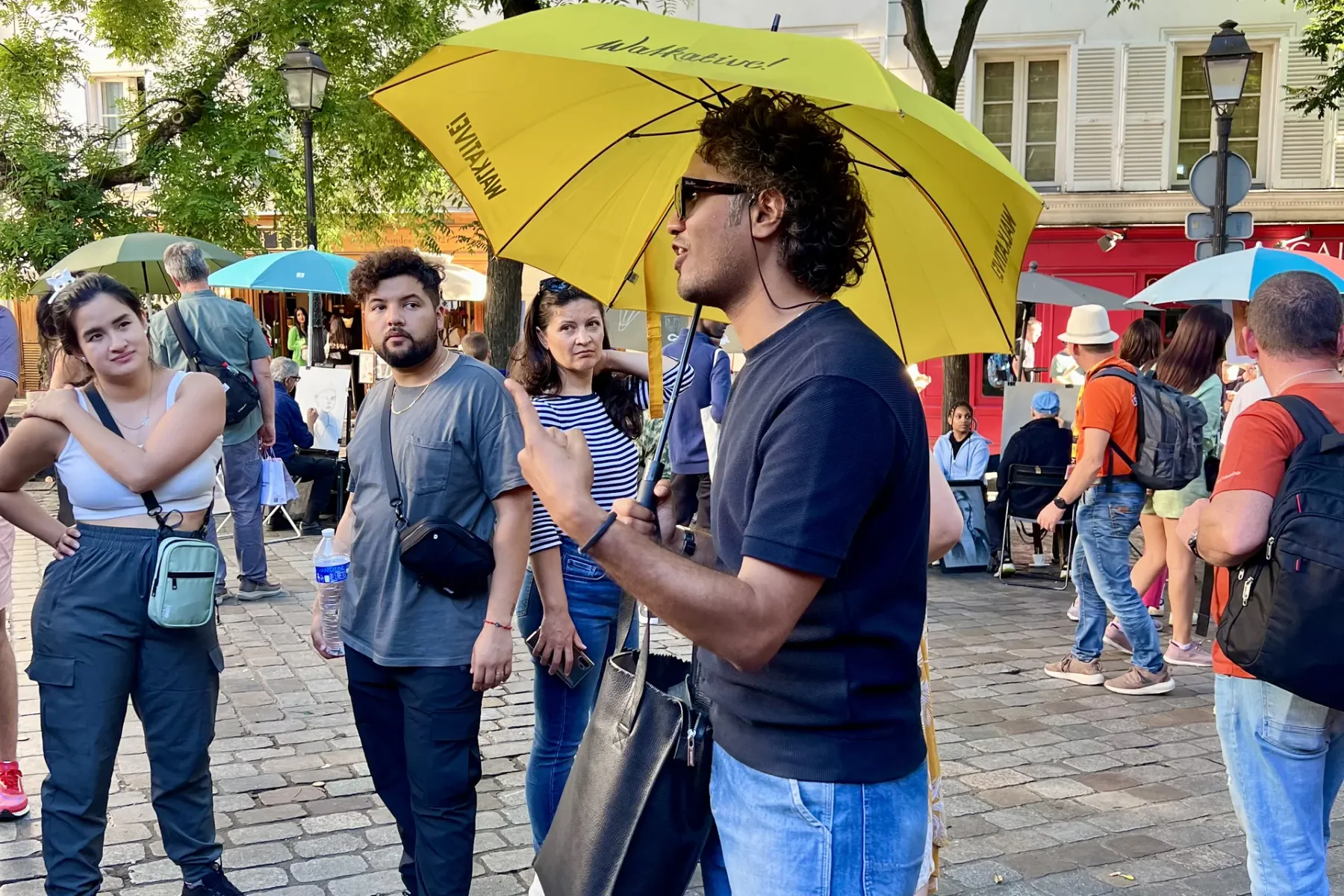 Ein Reiseführer unter einem gelben Walkative-Schirm spricht mit einer kleinen Touristengruppe auf einer Kopfsteinpflasters...