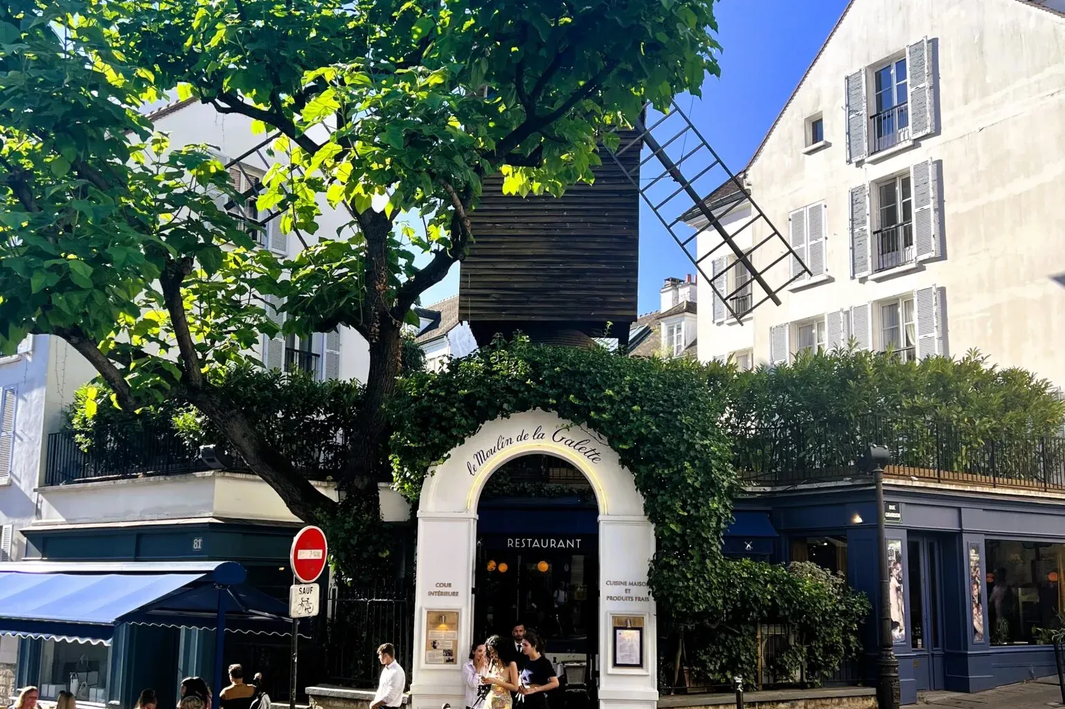 Restaurant Le Moulin de la Galette in Paris mit historischer Windmühlenstruktur, Außenterrasse, grünen Bäumen und Kopfstei...