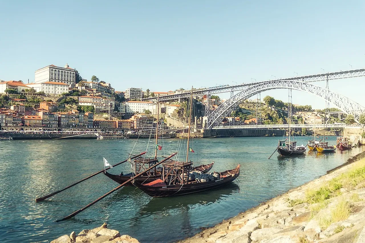 Traditionelle Rabelo-Boote auf dem Fluss Douro, mit bunten Gebäuden Portos und der Brücke Dom Luís I im Hintergrund.