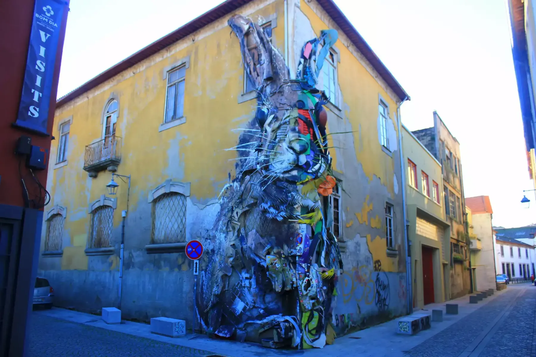Große Hasenskulptur aus recycelten Materialien an der Ecke eines gelben Gebäudes in Porto an einer Kopfsteinpflasterstraße.