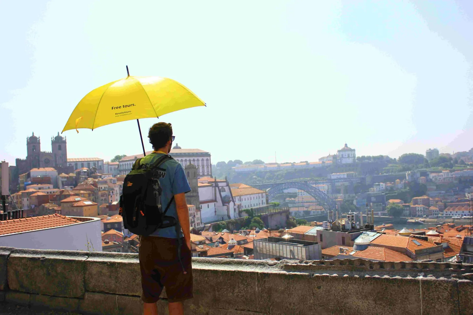 Person mit gelbem Regenschirm, von hinten gesehen, blickt auf Portos Skyline mit Dom Luís I Brücke und Kathedraltürmen.