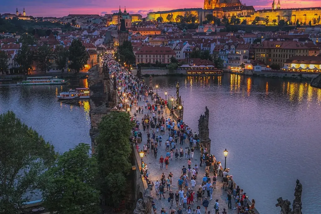 Puente de Carlos en Praga con muchos peatones al atardecer, el Castillo de Praga iluminado al fondo sobre el río Moldava.