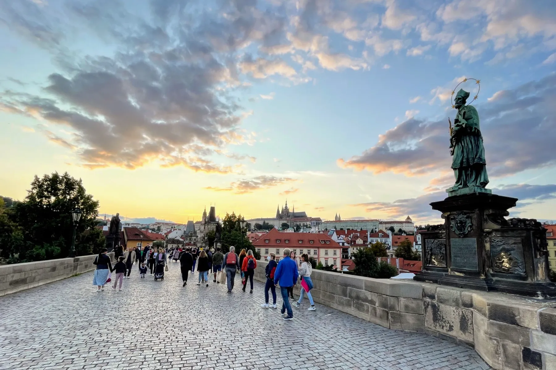 Puente de Carlos adoquinado con gente caminando al atardecer, estatua de San Juan Nepomuceno a la derecha, Castillo de Pra...