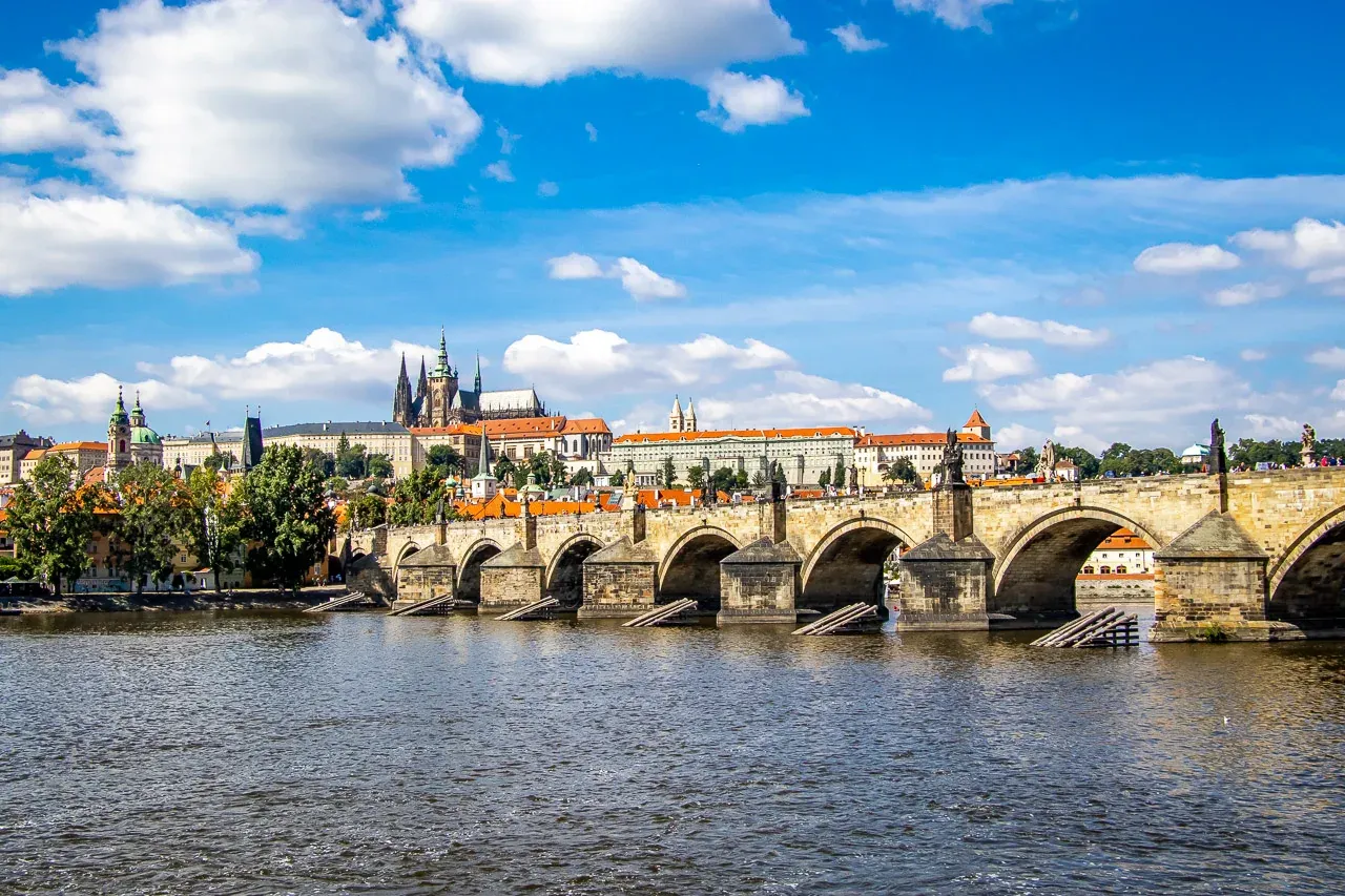 Vista del Puente de Carlos con estatuas y arcos sobre el río, el Castillo de Praga en la orilla izquierda bajo cielo azul ...