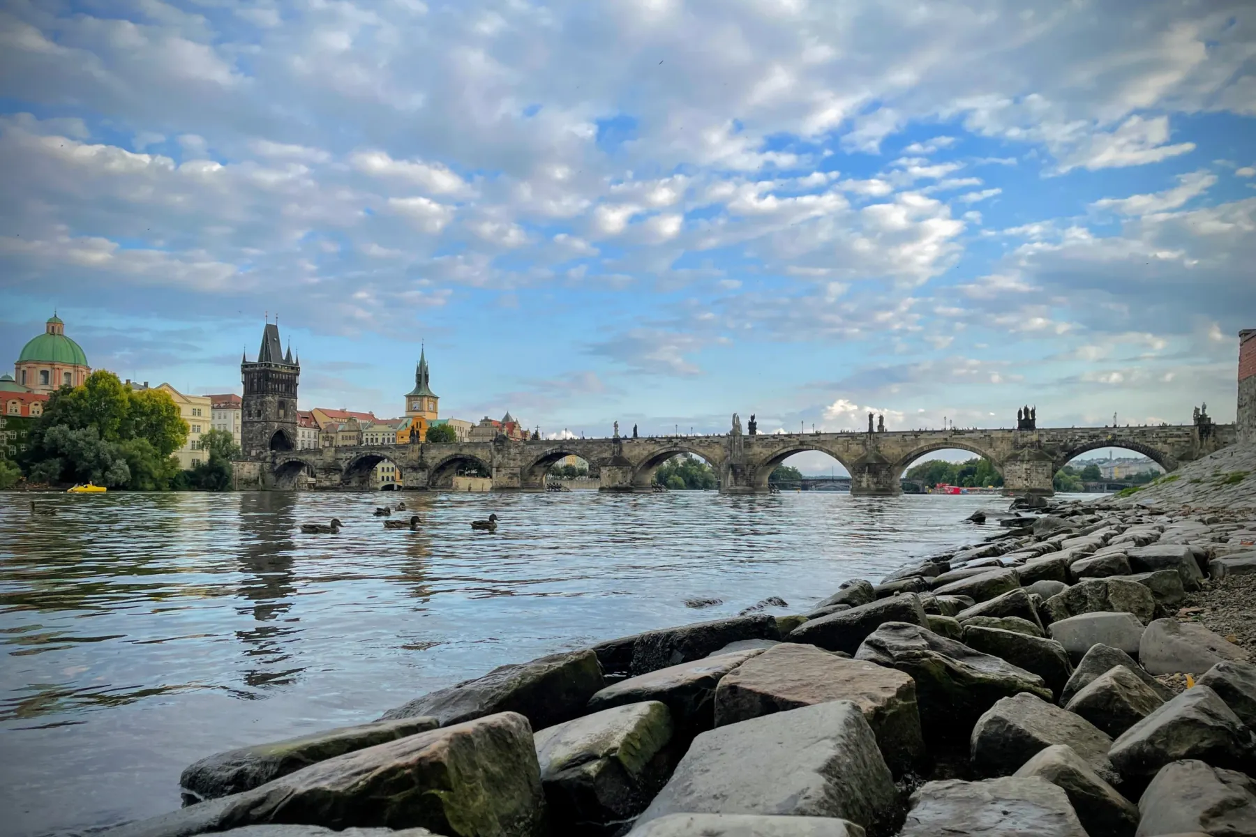 Vista del Puente de Carlos desde la orilla del río Moldava con patos, edificios históricos y cielo nublado.