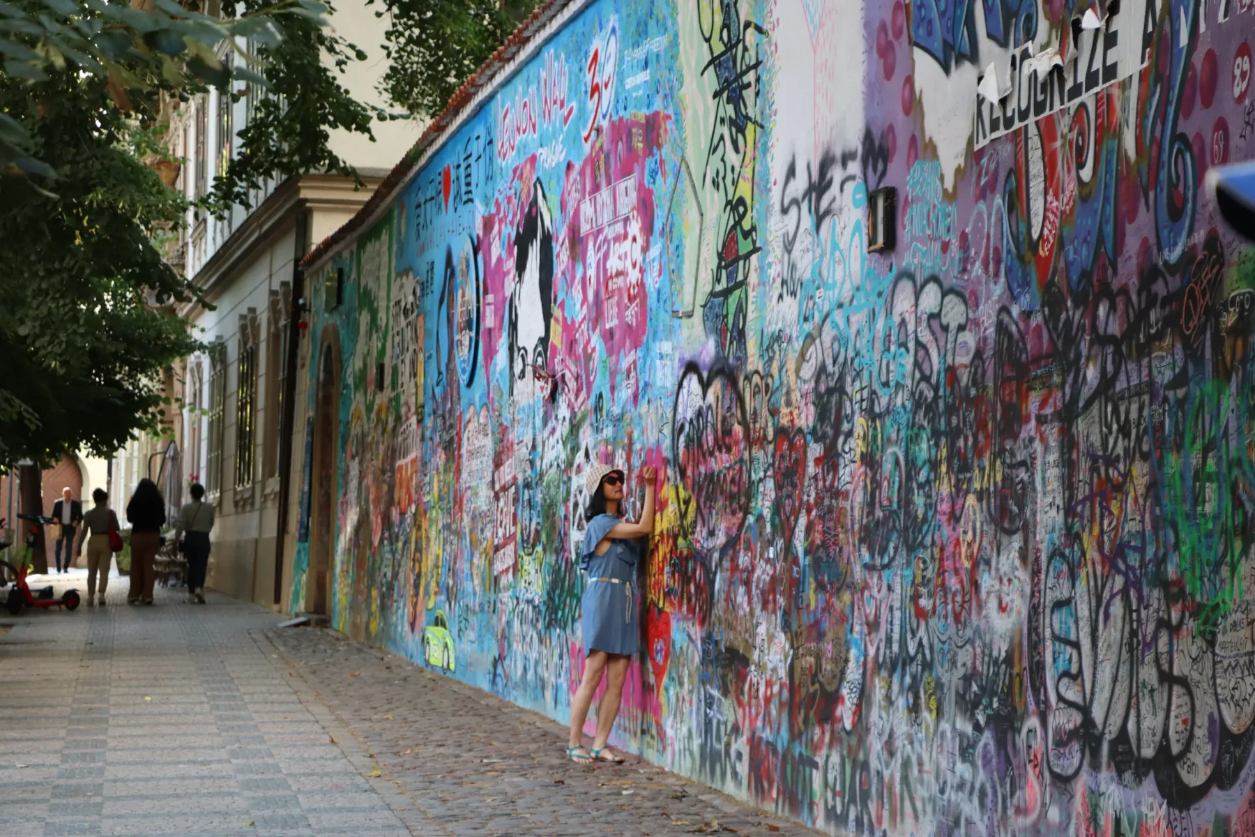 Una mujer con vestido azul y sombrero de paja junto al Muro de John Lennon en Praga, cubierto de grafitis coloridos.