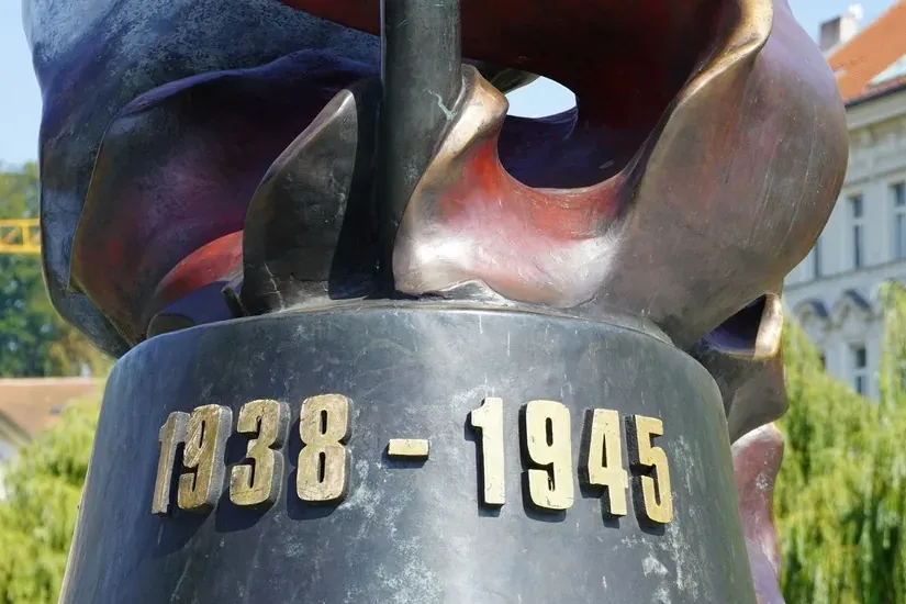 Primer plano de la base de un monumento de bronce con las fechas doradas '1938 - 1945' inscritas. Una torre de iglesia y á...
