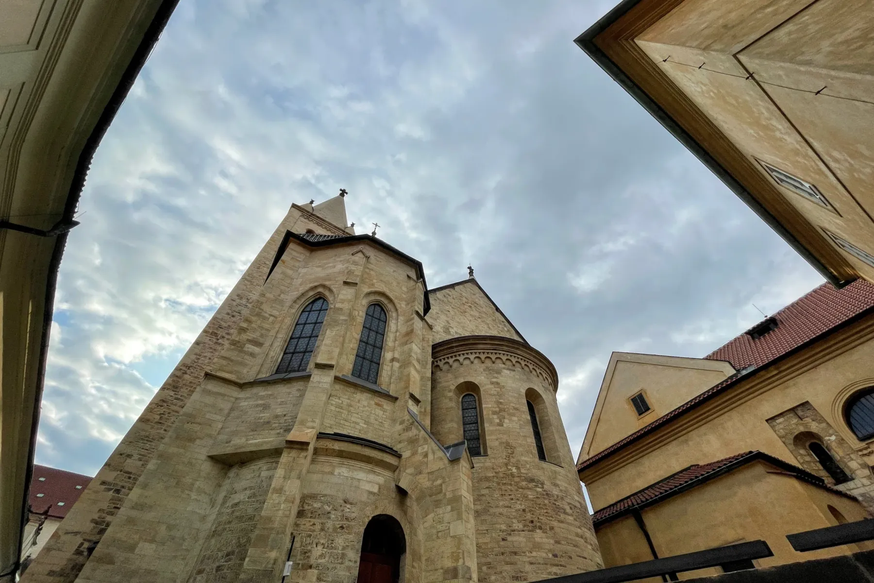 Edificio de iglesia de piedra con alta aguja y ventanas arqueadas, enmarcado por edificios adyacentes y cielo nublado.