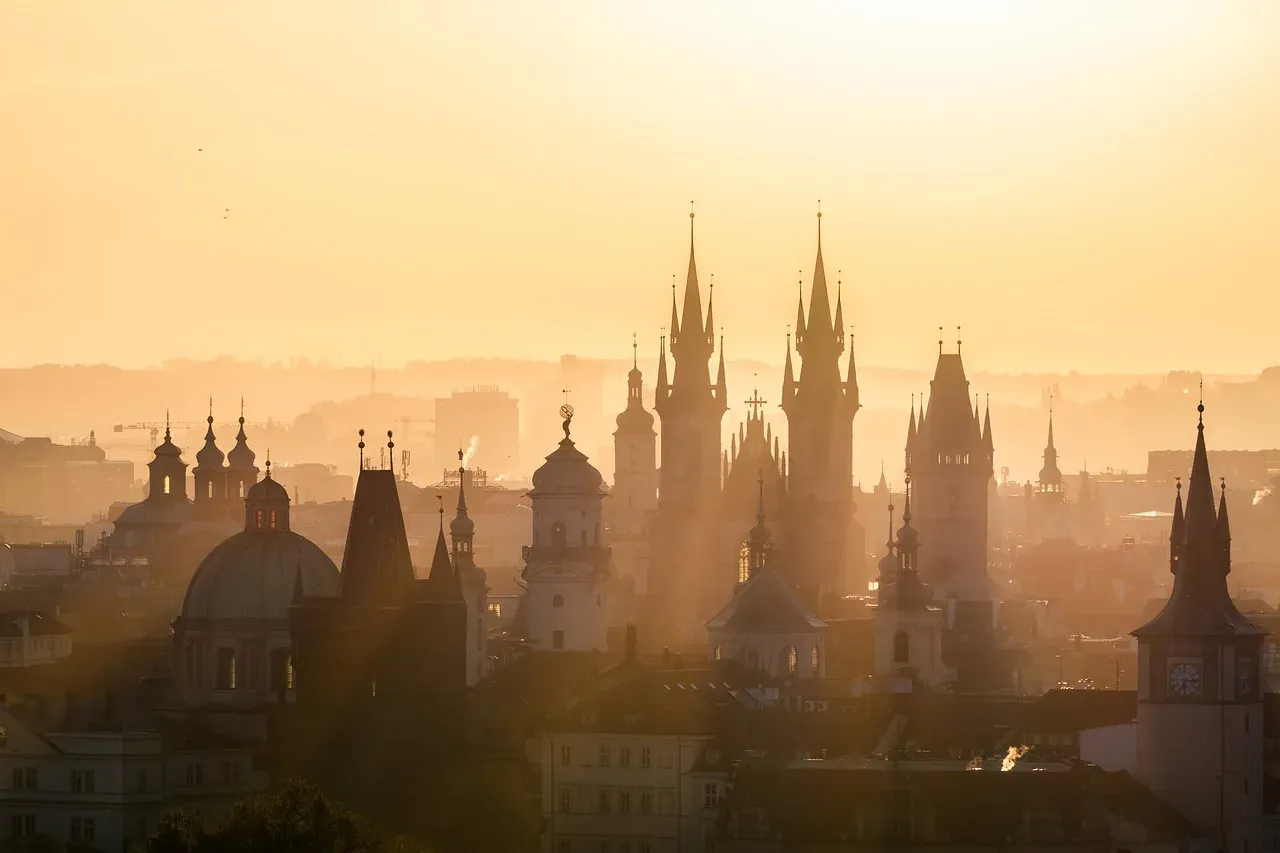 Denso paisaje urbano de Praga al atardecer, mostrando cúpulas, agujas y torres históricas contra un cielo brumoso y anaran...