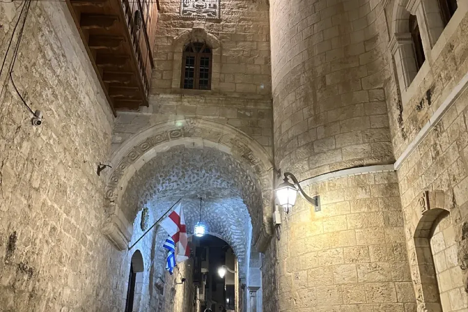 Callejón estrecho de piedra de noche con un arco, escalones, dos motos y un edificio con cruz y balcón de madera.