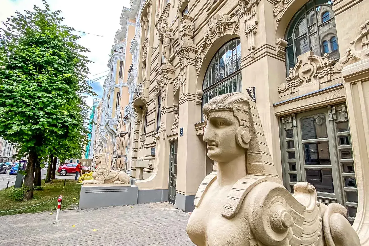 Helle Sphinx-Statue mit menschlichem Kopf und geflügeltem Körper auf einem Sockel, vor einem verzierten Jugendstilgebäude ...