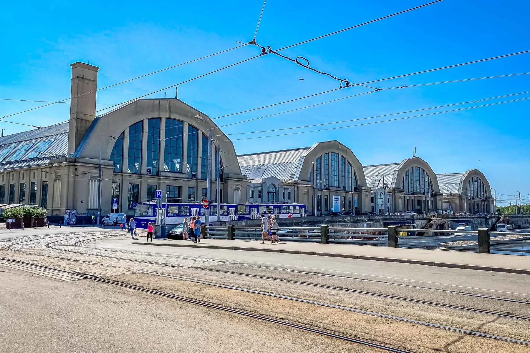 Fünf große Markthallen mit gewölbten Dächern und Fenstern, Straßenbahnschienen im Vordergrund, Menschen auf einer Straße i...