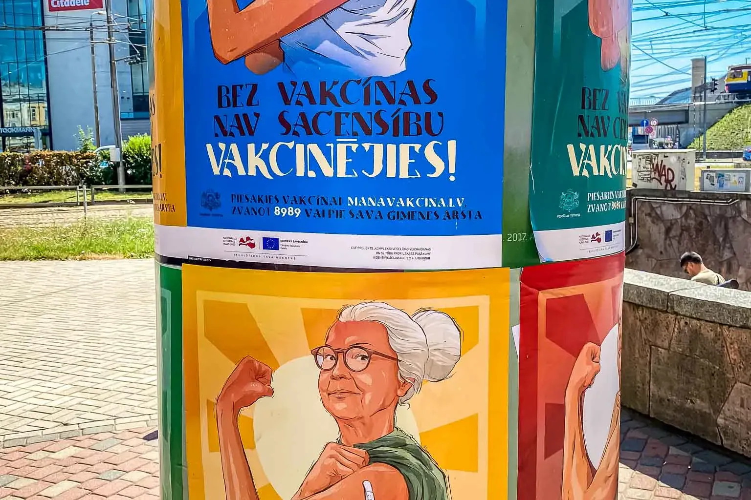Zylindrischer Litfaßsäule mit Plakaten einer Impfkampagne in Riga, darunter eine Tennis spielende Frau und eine ältere Fra...