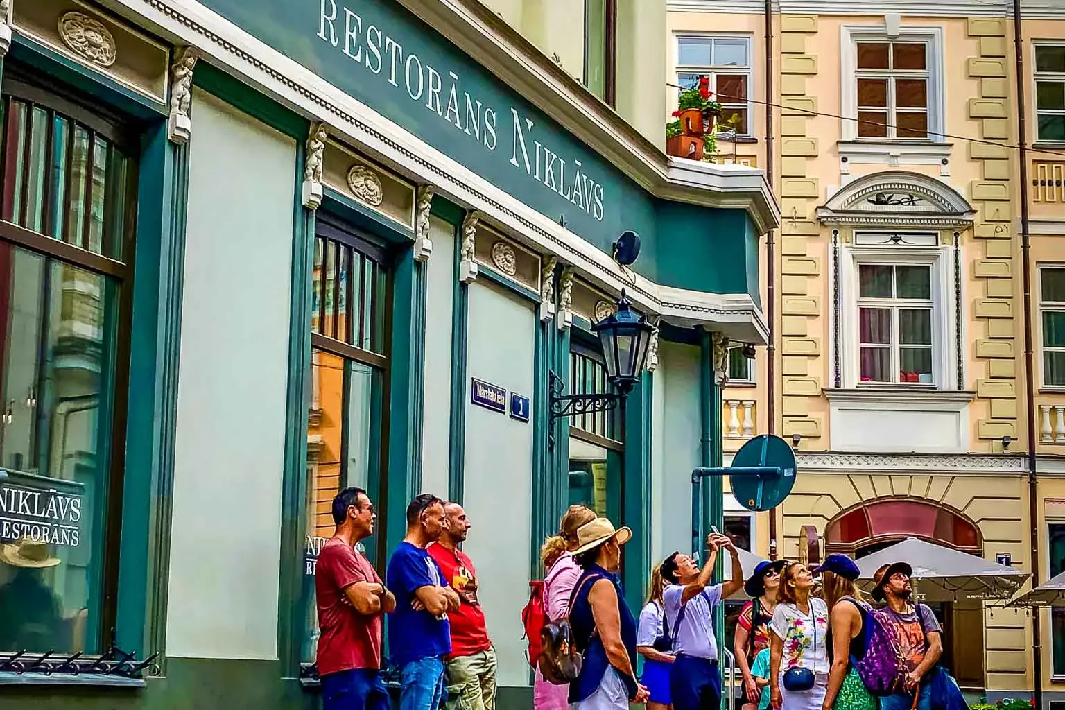Eine Gruppe Menschen blickt zu der Fassade eines hellgrünen Gebäudes mit der Aufschrift „RESTORĀNS NIKLĀVS“ auf. Historisc...