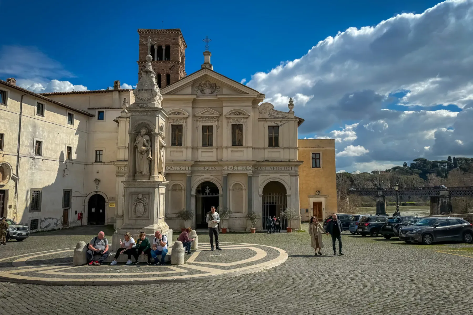 Blick auf die Basilika San Bartolomeo und ein Denkmal auf dem Platz der Tiberinsel, mit Menschen und Autos unter bewölktem...
