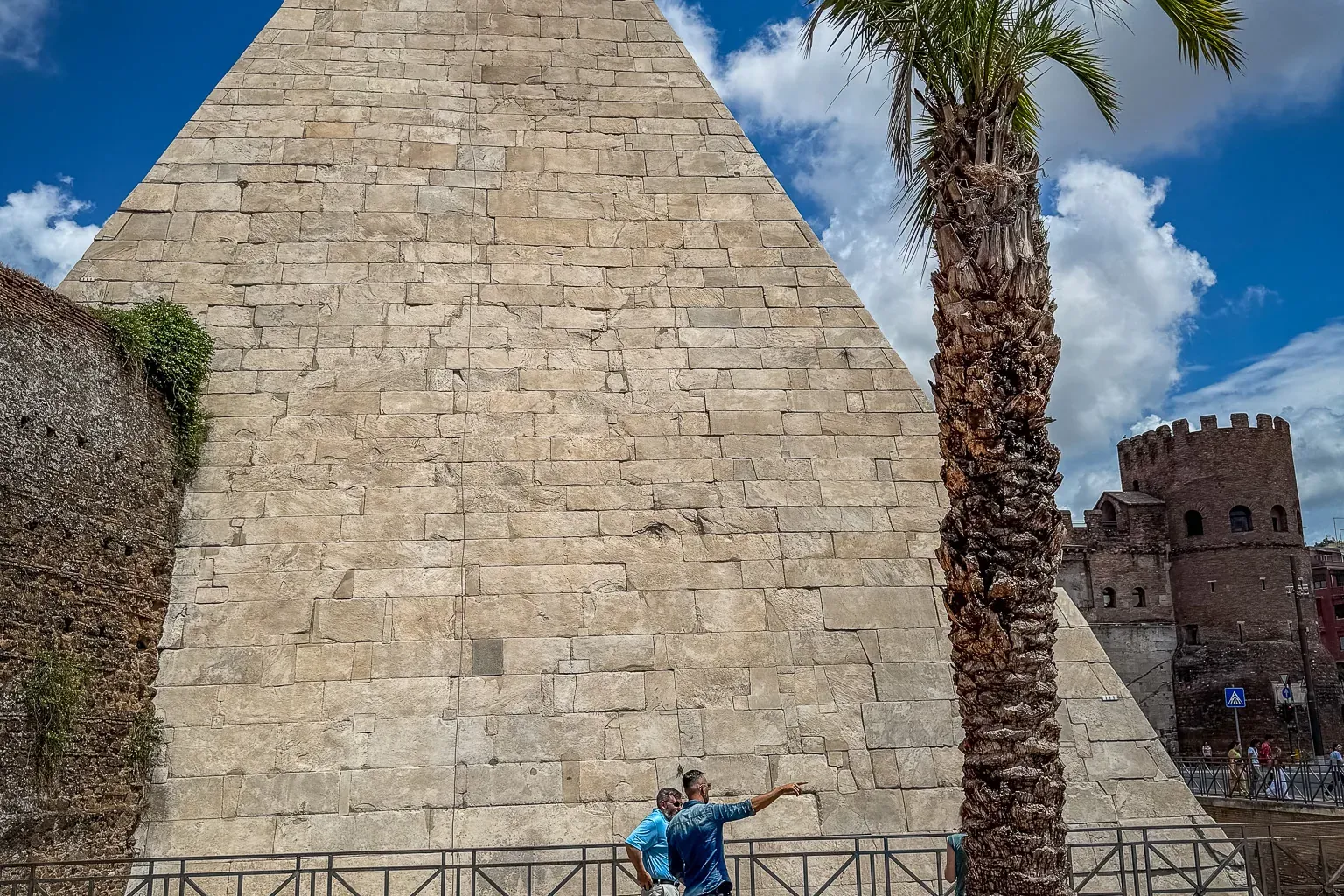 Die Cestius-Pyramide in Rom, ein großes Steinbauwerk, mit einer hohen Palme und zwei Touristen davor.