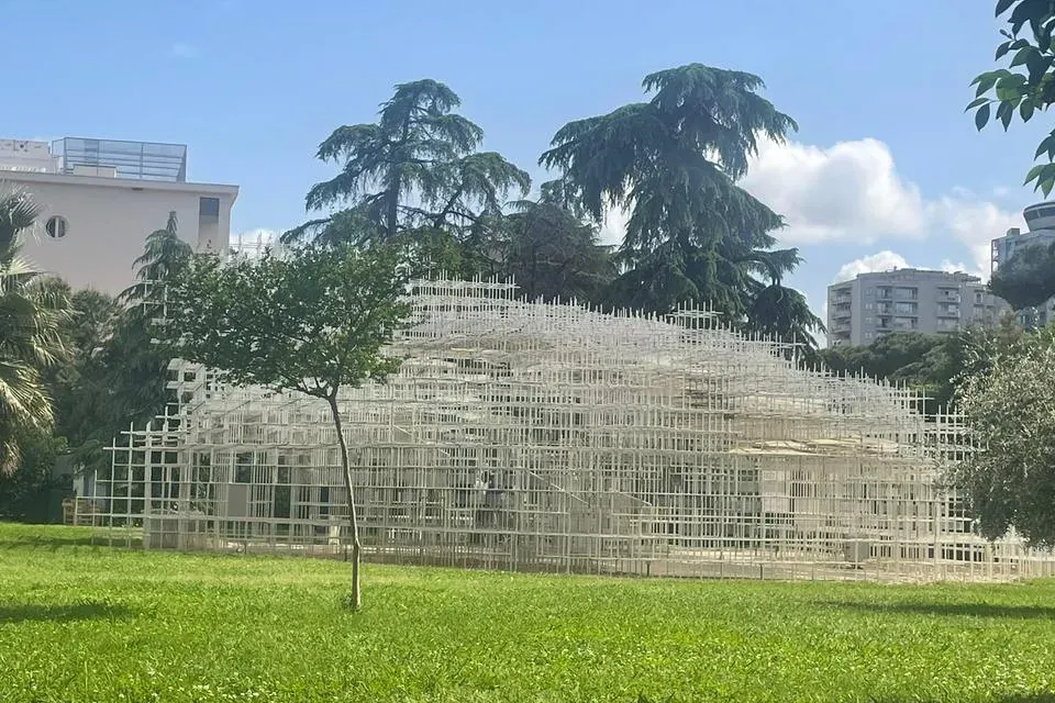 Duża, biała, siatkowa instalacja artystyczna w zielonym parku pod błękitnym niebem z chmurami. W tle widoczne drzewa i bud...