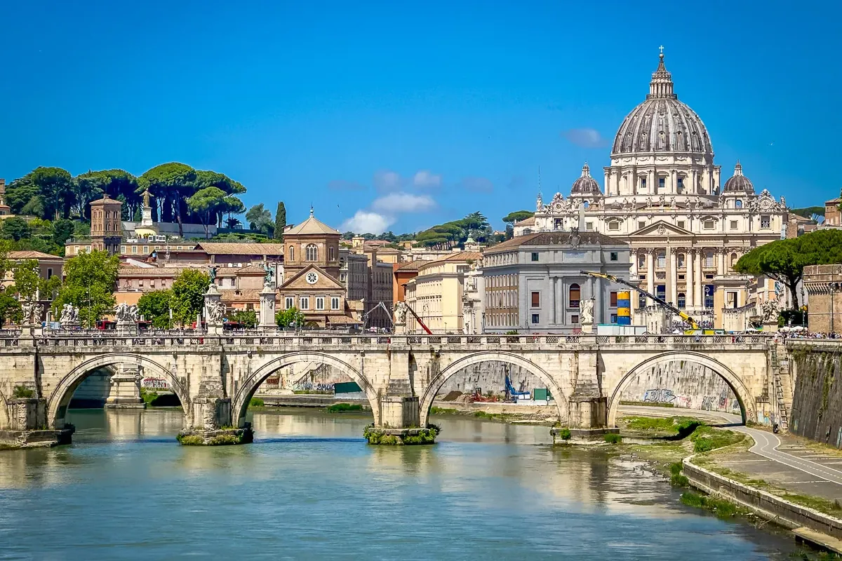 Engelsbrücke über den Tiber, mit Kuppel des Petersdoms und römischen Gebäuden unter klarem blauem Himmel.