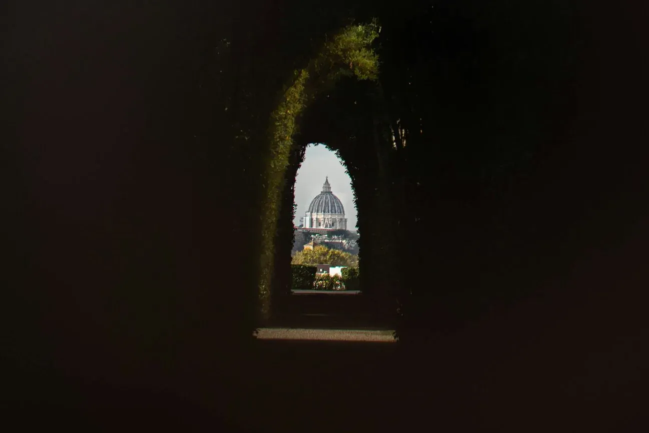 Die Kuppel des Petersdoms, durch eine kleine, dunkle, schlüssellochförmige Öffnung in einer Hecke oder Mauer mit Grünpflan...
