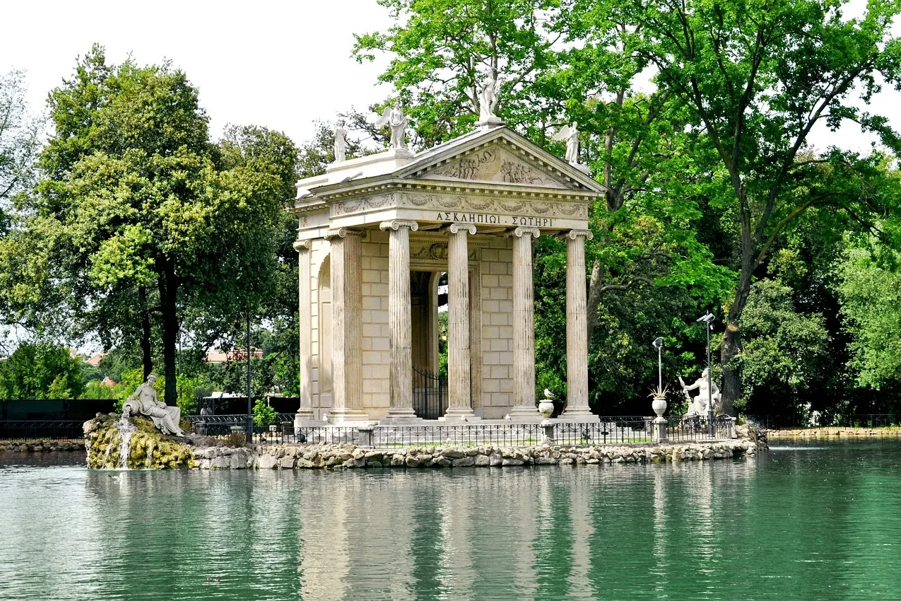 Klassischer Tempel auf einer kleinen Insel in einem ruhigen Teich, umgeben von üppig grünen Bäumen und Skulpturen.