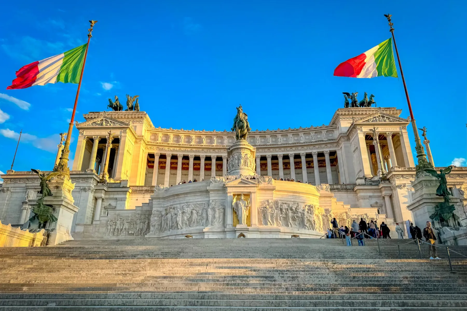 Das Nationaldenkmal Vittoriano in Rom, ein weißes Marmormonument mit Säulen, Statuen und italienischen Flaggen, bei blauem...