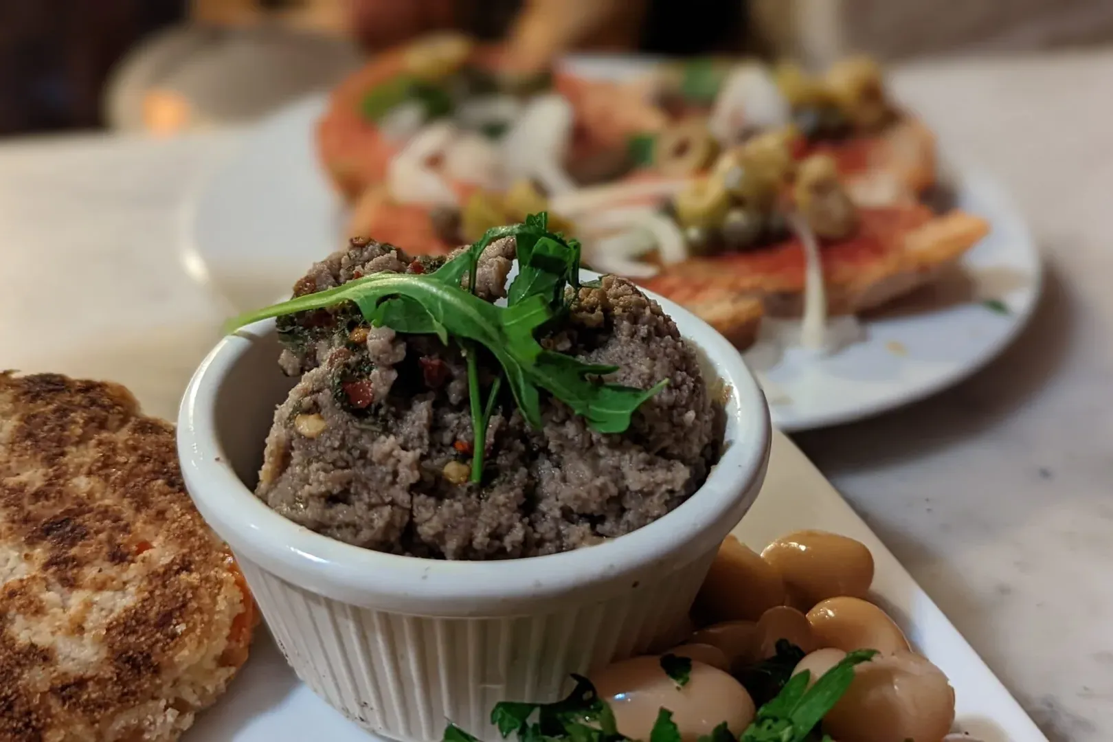 Primer plano de una cazuela blanca con paté marrón y rúcula, una tortita redonda y alubias blancas con hierbas. Bruschetta...
